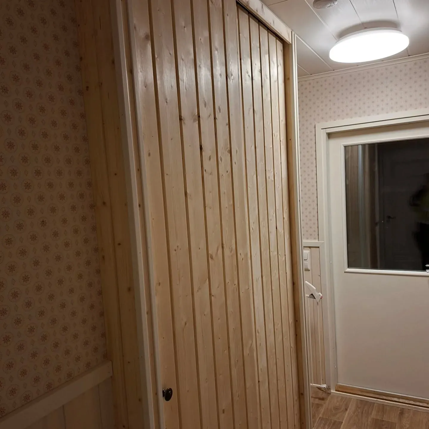 Sisäkuva vaaleasta puupaneeliseinästä tai kaapista vasemmalla. Oikealla on valkoinen ovi, jossa on ikkuna. Seinät on tapetoitu vaalealla tapetilla, jossa on pieniä ruskeita kuvioita. Katossa on valkoinen paneeli, kirkas pyöreä kattovalo ja palovaroitin. Lattia on vaaleanruskeaa puuta. Oven edessä näkyy valkoinen turvaportti.