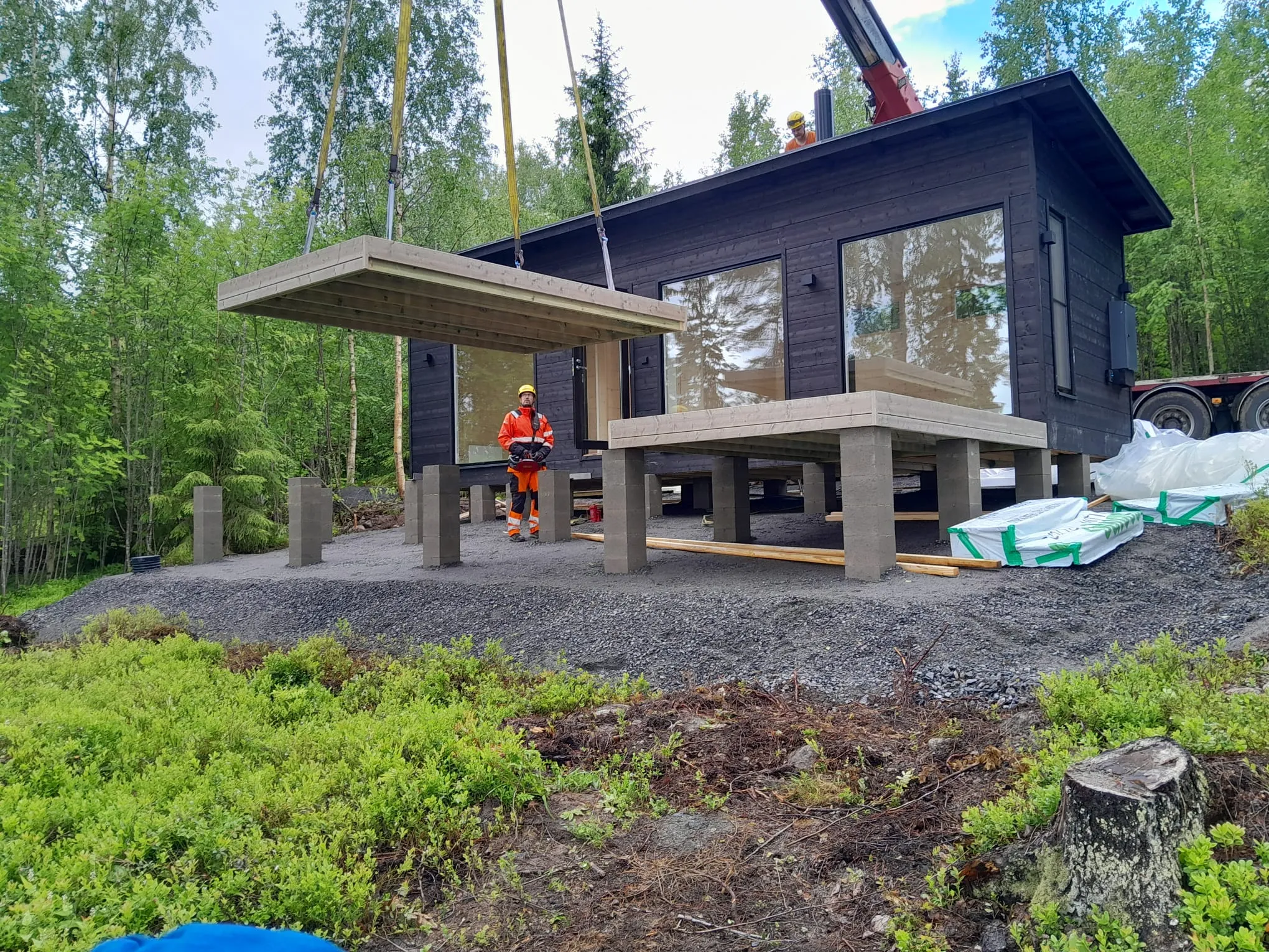 Tumma, moderni talo rakenteilla metsäisessä ympäristössä. Nosturi nostaa suurta puuterassiosaa kohti taloa. Yksi työntekijä oranssissa huomiopuvussa ja keltaisessa kypärässä ohjaa nostoa maasta käsin, ja toinen työntekijä on talon katolla. Talo on pystytetty betonipilareiden päälle harmaalle soralle. Taustalla on tiheää vihreää metsää ja etualalla aluskasvillisuutta sekä puun kanto. Valo on tasainen ja pilvinen.