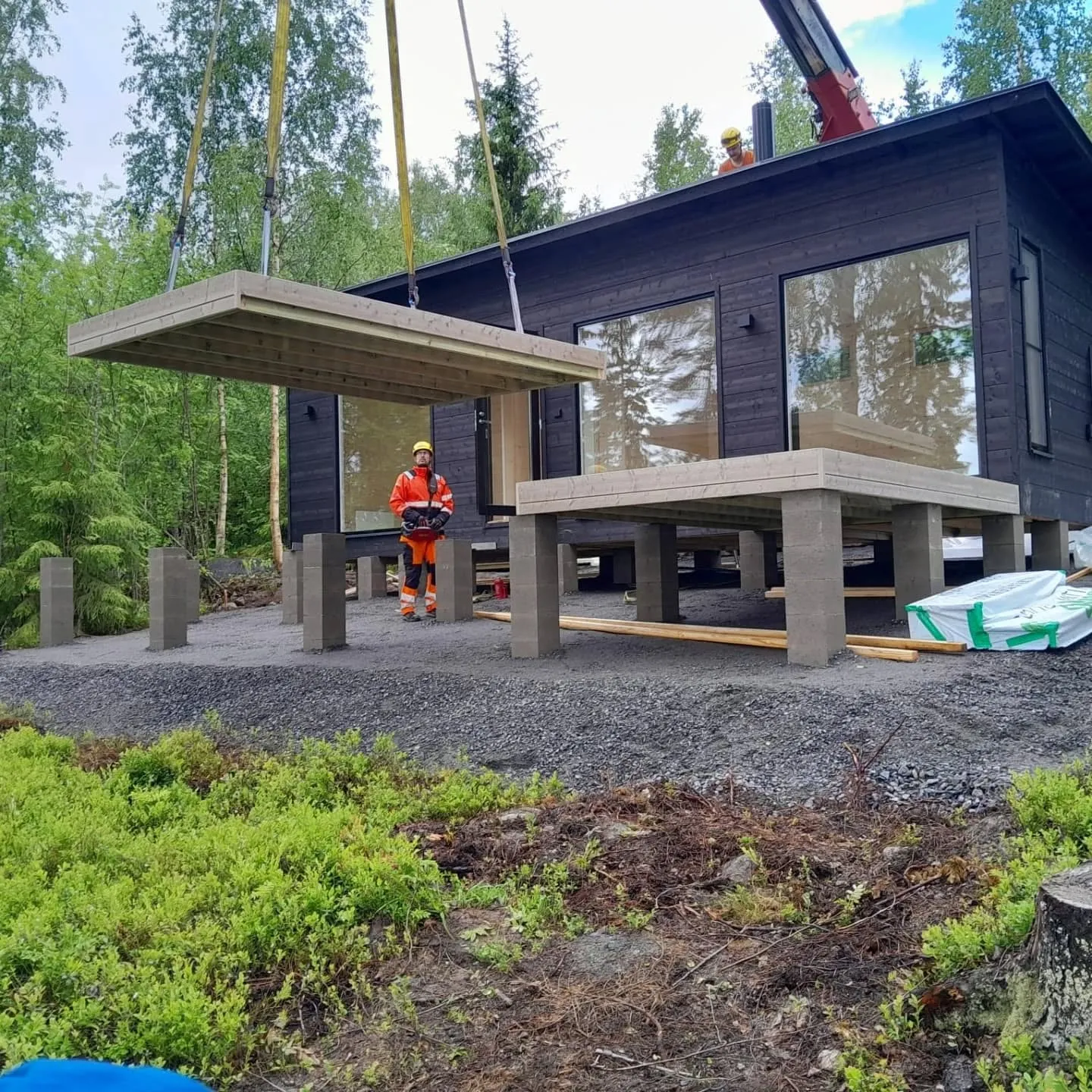 Kuva esittää rakennustyömaata metsäisessä ympäristössä. Nosturi laskee suurta puupaneelia tumman, puisen talon viereen, joka seisoo betonipilareiden päällä. Yksi työntekijä oranssissa työasussa ja keltaisessa kypärässä ohjaa paneelia maasta käsin. Toinen työntekijä on talon katolla. Maassa on soraa ja vihreää aluskasvillisuutta. Valaistus on kirkas, pilvinen päivä.