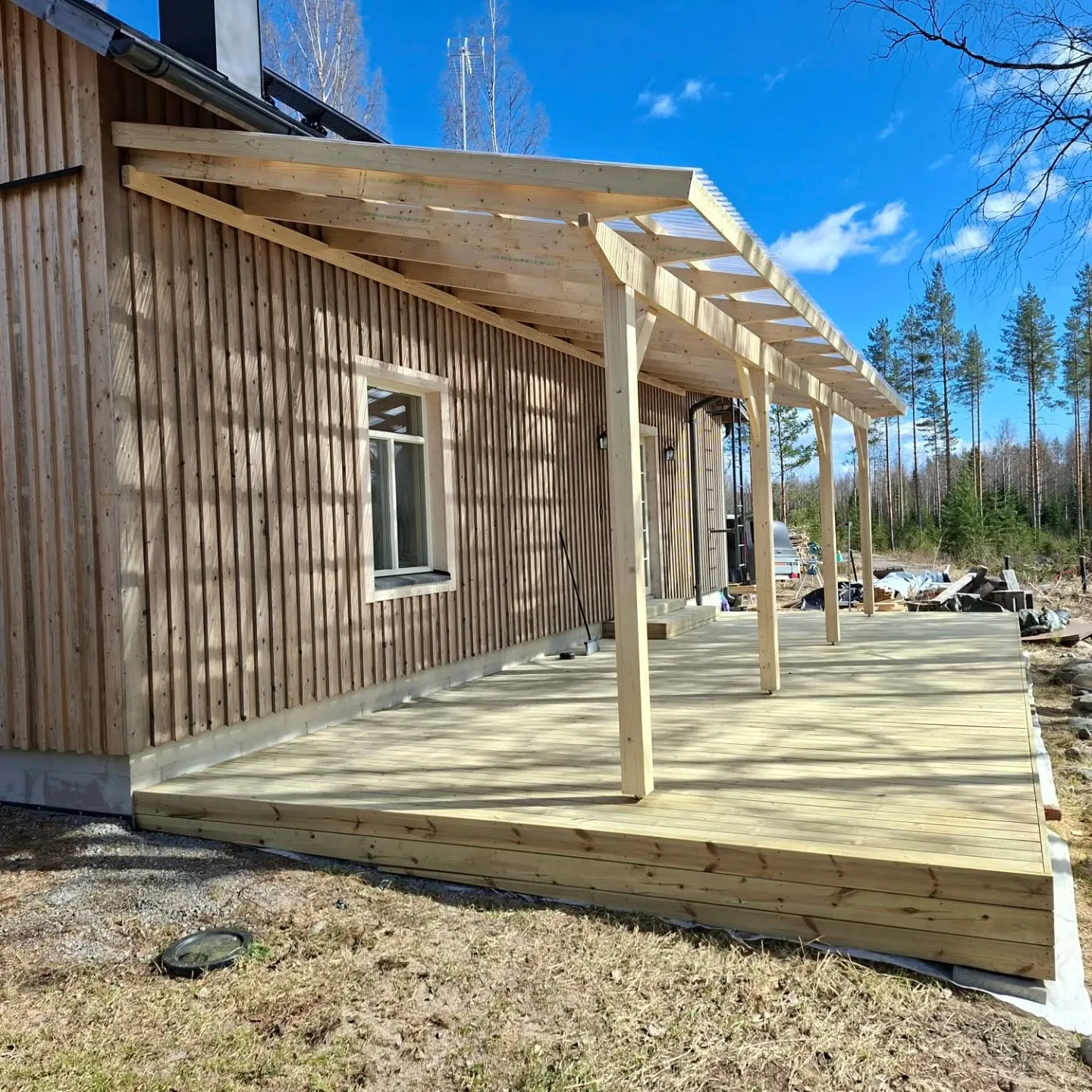 Vaaleanruskea puutalo pystysuoralla puuverhouksella ja valkoisella ikkunalla. Talon edessä on uusi, vaaleasta puusta rakennettu terassi, jota peittää läpinäkyvä aaltomuovikatto. Terassia tukevat puupylväät. Taustalla on metsää ja kirkas sininen taivas valkoisine pilvineen. Aurinkoinen päivä luo varjoja terassille ja talon seinään.