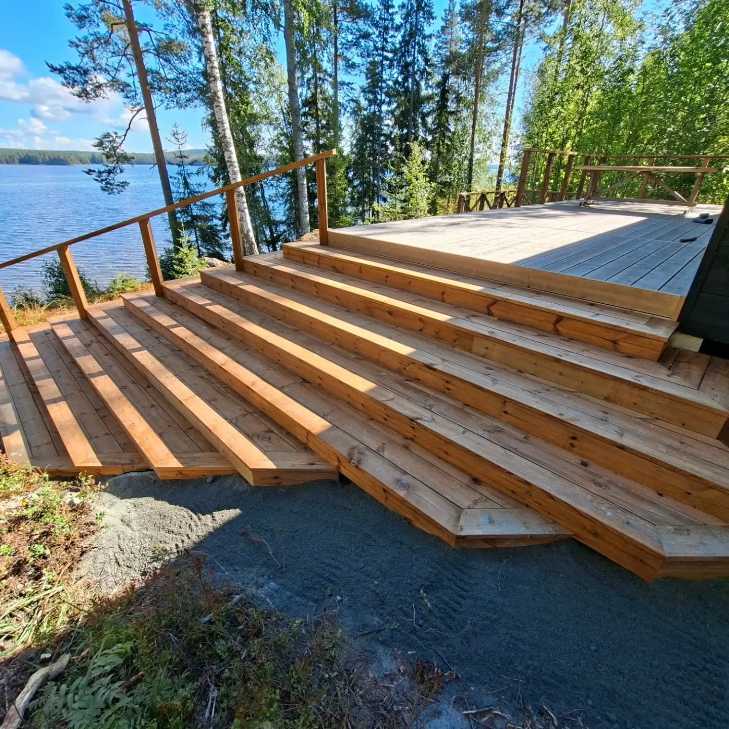 Aurinkoinen kuva puuterassista ja leveistä puuportaista, jotka laskeutuvat kohti sinistä järveä. Vasemmalla on puinen kaide. Taustalla näkyy vihreää metsää ja kirkkaan sininen taivas. Maaperä portaiden juurella on harmaata ja siinä on kasvillisuutta. Auringonvalo luo pitkiä varjoja portaille ja terassille.