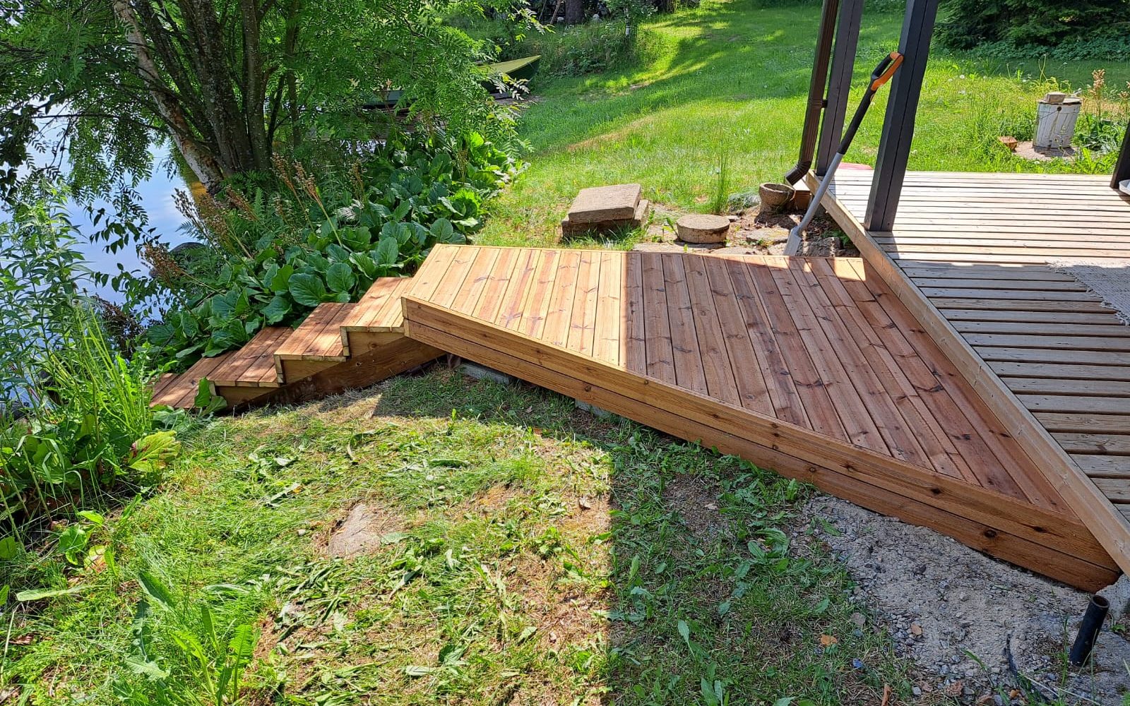 Aurinkoinen kuva puuterassista ja portaista, jotka laskeutuvat alas veden äärelle. Terassin puupinta on vaaleanruskea, osittain auringossa ja osittain varjossa. Portaiden ympärillä on rehevää vihreää ruohoa ja kasvillisuutta. Vasemmalla näkyy sininen vesi, ja taustalla on tiheää puustoa. Terassin vieressä maassa on muutamia betonilaattoja ja työkalu nojaa pylvästä vasten.