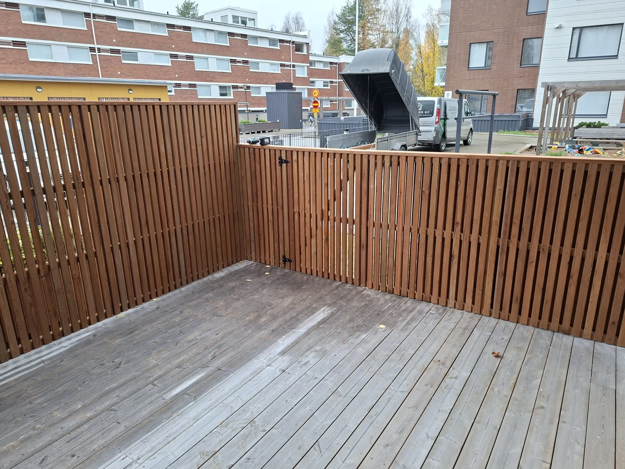 Kuvassa on ulkoterassi, jonka lattia on harmaantunutta puuta ja jolla on muutama pudonnut lehti. Terassia rajaa ruskea pystylauta-aita, jossa on musta portin salpa. Taustalla näkyy tiilirakennuksia valkoisilla ikkunoilla, keltainen rakennus ja valkoinen rakennus. Keskellä on katu, jolla on valkoinen pakettiauto ja musta peräkärry lava ylhäällä. Päivä on pilvinen ja valaistus tasainen.