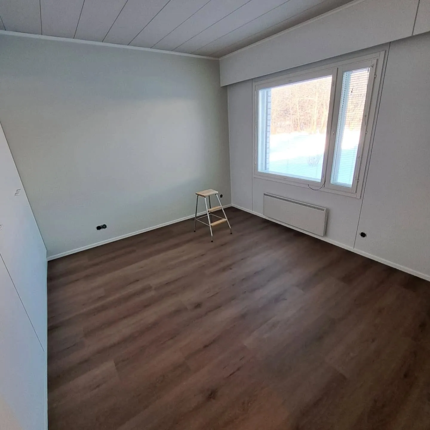 Tyhjä huone, jossa on vaaleanharmaat seinät, valkoinen paneelikatto ja tummanruskea laminaattilattia. Oikealla seinällä on suuri ikkuna, josta näkyy lumista metsämaisemaa. Ikkunan alla on valkoinen patteri. Huoneen keskellä on pieni puinen askeljakkara. Huone on valaistu luonnonvalolla.