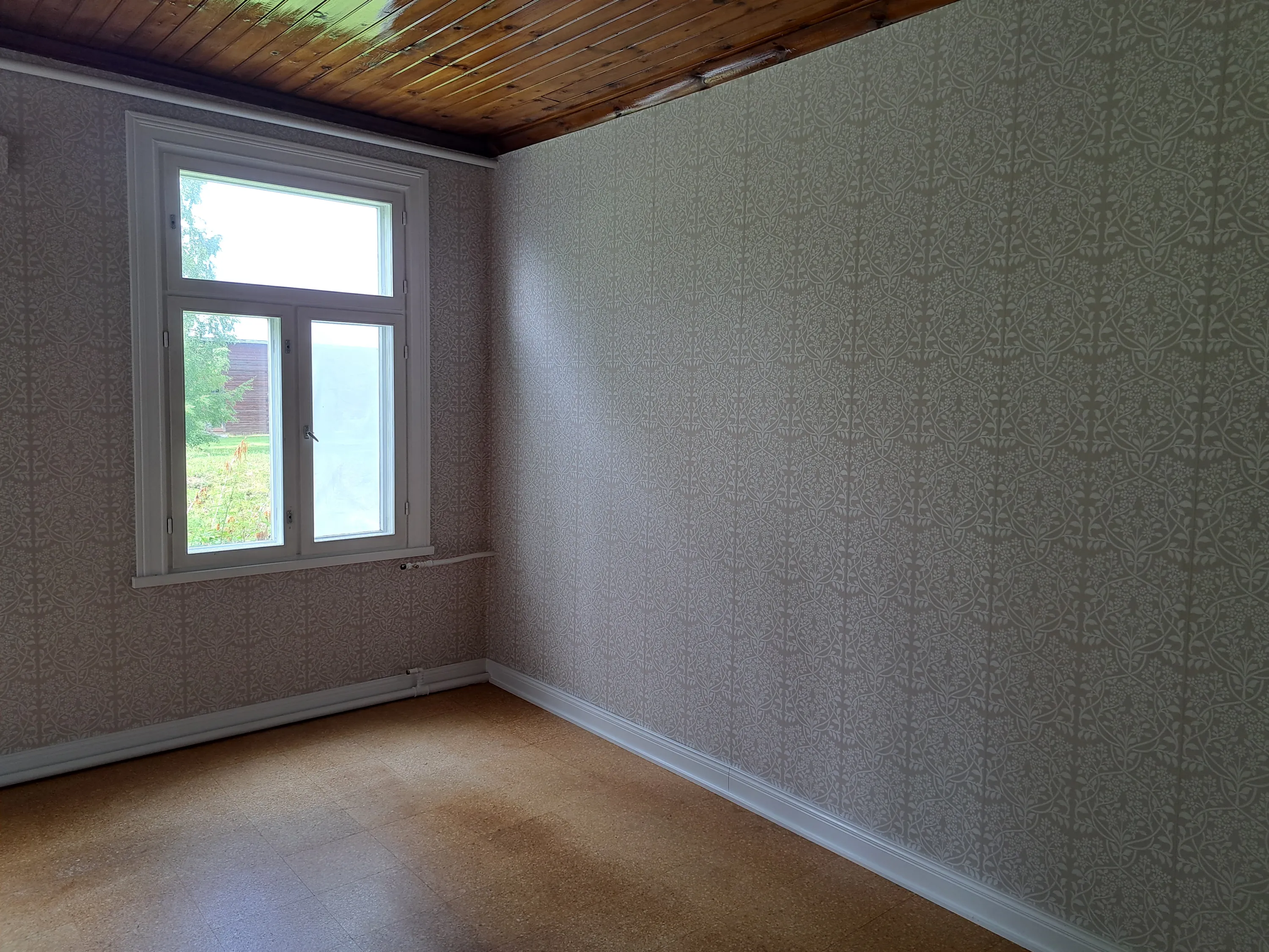 Tyhjä huone, jossa on vaaleanharmaa kasviaiheinen tapetti seinillä. Vasemmalla on valkoinen ikkuna, josta avautuu näkymä vihreälle pihalle ja puurakennukselle. Katossa on kiiltävä, punertavanruskea puupaneeli. Lattia on vaaleanruskeaa, laattamaisen näköistä materiaalia, ja siihen lankeaa varjo. Valkoiset jalkalistat kiertävät huonetta, ja ikkunan alla on valkoisia putkia.