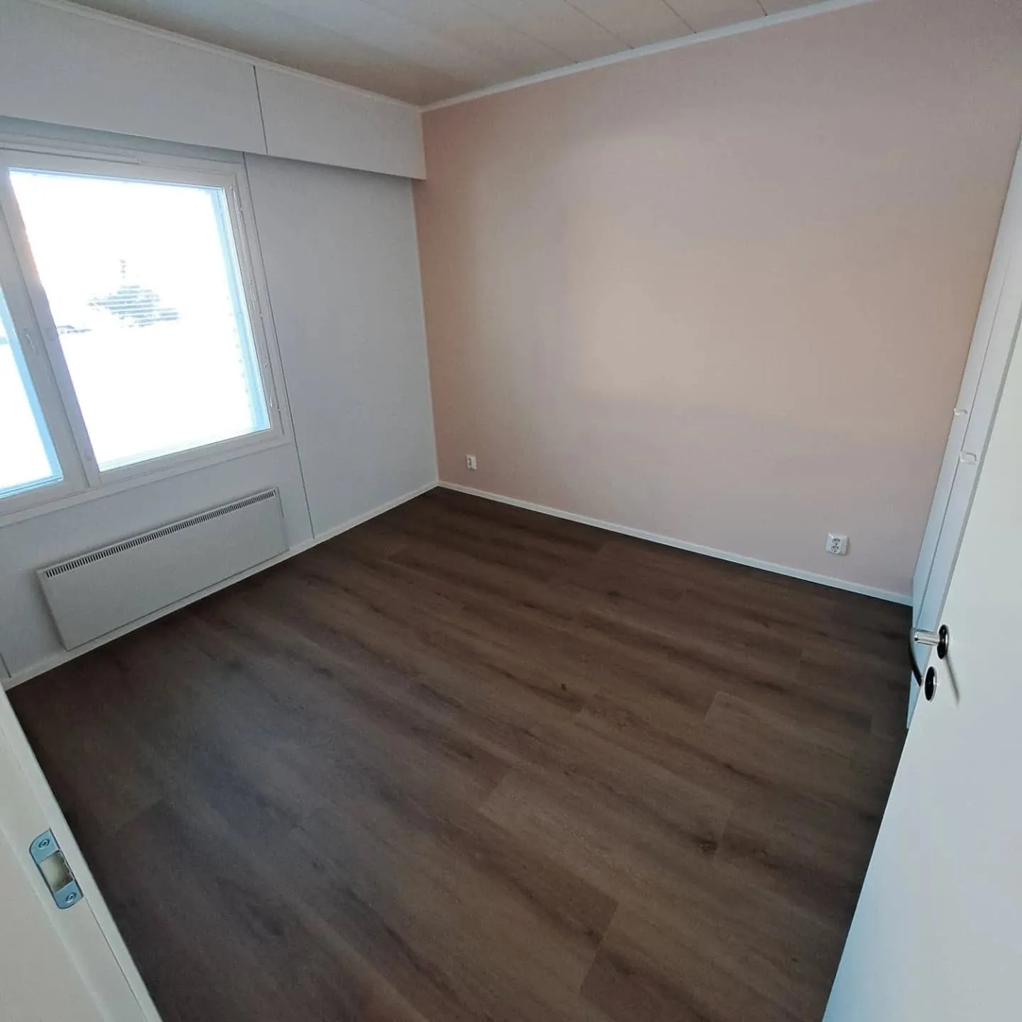 Tyhjä huone, jossa on tummanruskea laminaattilattia ja valkoinen katto. Vasemmalla seinällä on suuri ikkuna, jonka alla on valkoinen lämpöpatteri. Oikeanpuoleinen seinä on vaaleanpunainen, ja muut seinät ovat valkoisia. Huone on valoisa luonnonvalon ansiosta. Oikealla näkyy valkoisen oven karmi.