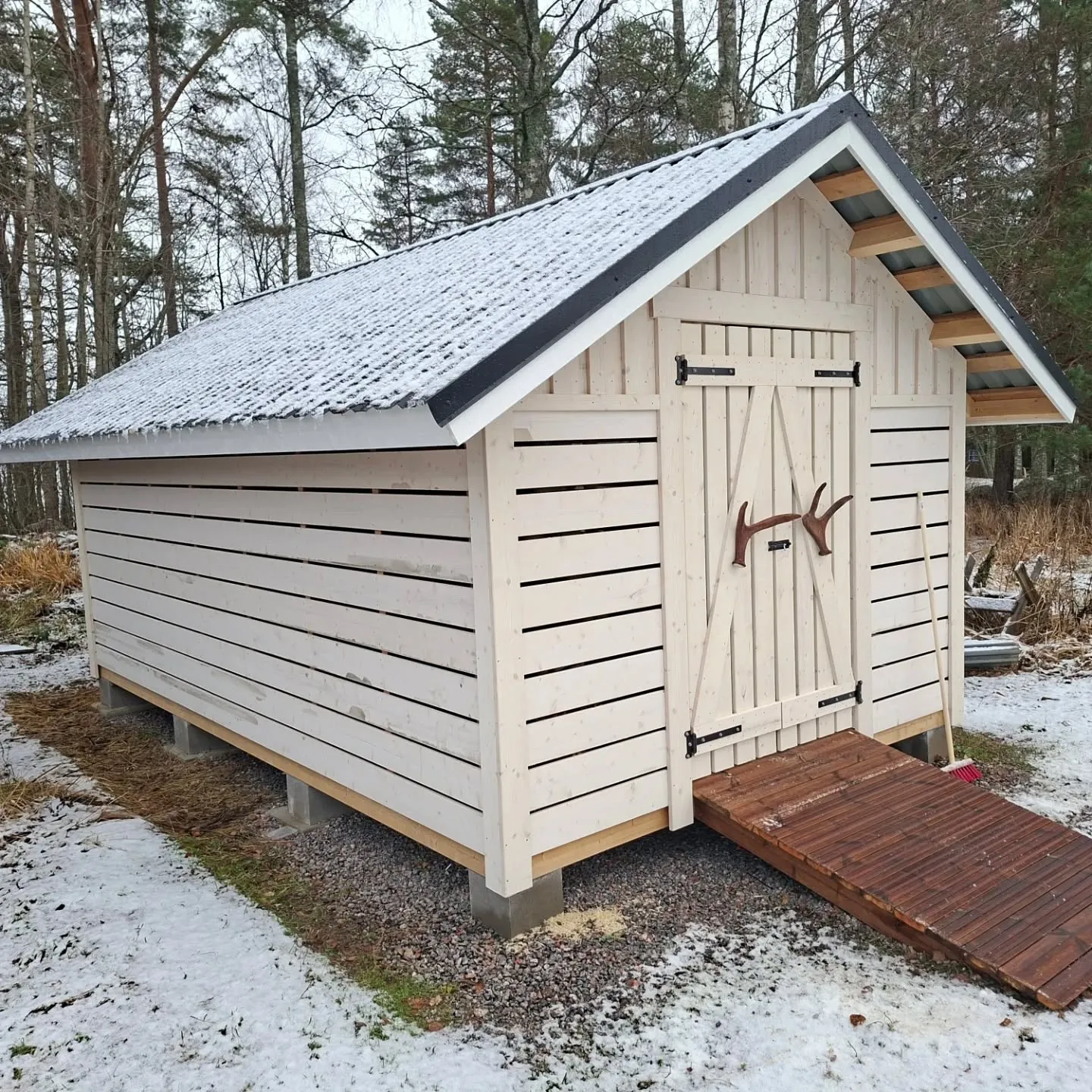 Vaalea puinen varastorakennus seisoo lumisessa maisemassa. Sen tummanharmaa aaltopeltikatto on ohuen lumikerroksen peitossa. Rakennuksen etupuolella on pariovet, joissa on mustat saranat ja ruskeat sarvenmuotoiset kahvat. Oven edessä on tummanruskea puinen luiska. Rakennus on perustettu harmaille betoniharkoille. Maassa on lunta, soraa ja kuivaa ruohoa. Taustalla näkyy puita pilvisen taivaan alla.